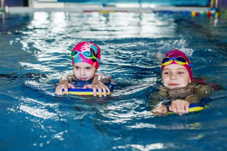 Children learn to swim with board in pool under guidance of coach. Swimming lesson. Active kids are playing in water. Girls in goggles and swimming cap